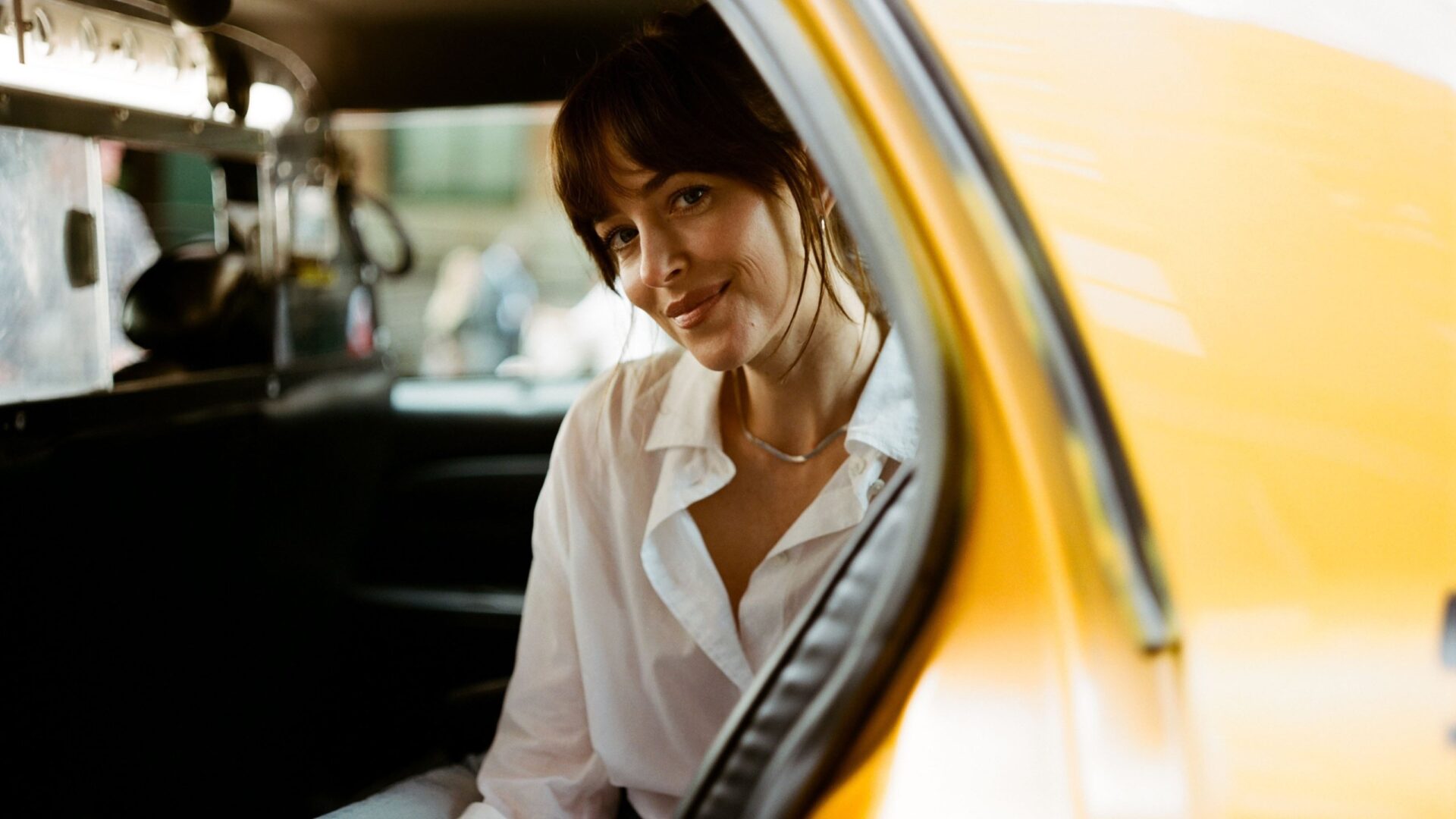 Auf dem Bild sitzt Dakota Johnson in einem Taxi und lächelt in die Kamera