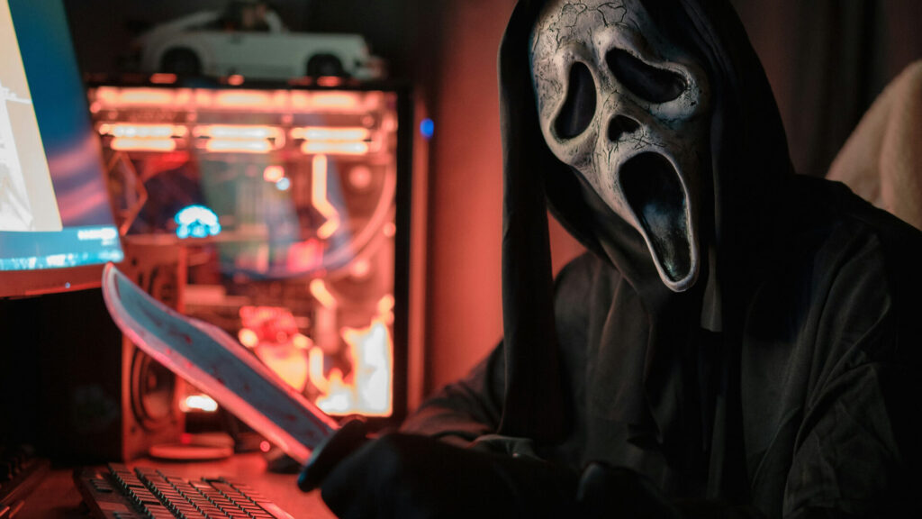 Eine Person sitzt in einem abgedunkelten Raum vor einem Computer und trägt ein vollständiges Ghostface-Kostüm aus der „Scream“-Filmreihe, inklusive weißer Maske und schwarzem Umhang. Die Person hält ein großes Messer bedrohlich in der Hand. Der PC-Tower leuchtet auffällig in Rot und Blau; darauf steht ein kleines Spielzeugauto. Links im Bild ist ein Monitor teilweise sichtbar. Die Szene wirkt bewusst inszeniert und erzeugt eine düstere, unheimliche Stimmung mit Anklängen an Cyber-Horror. | Themenübersicht Oktober 2025