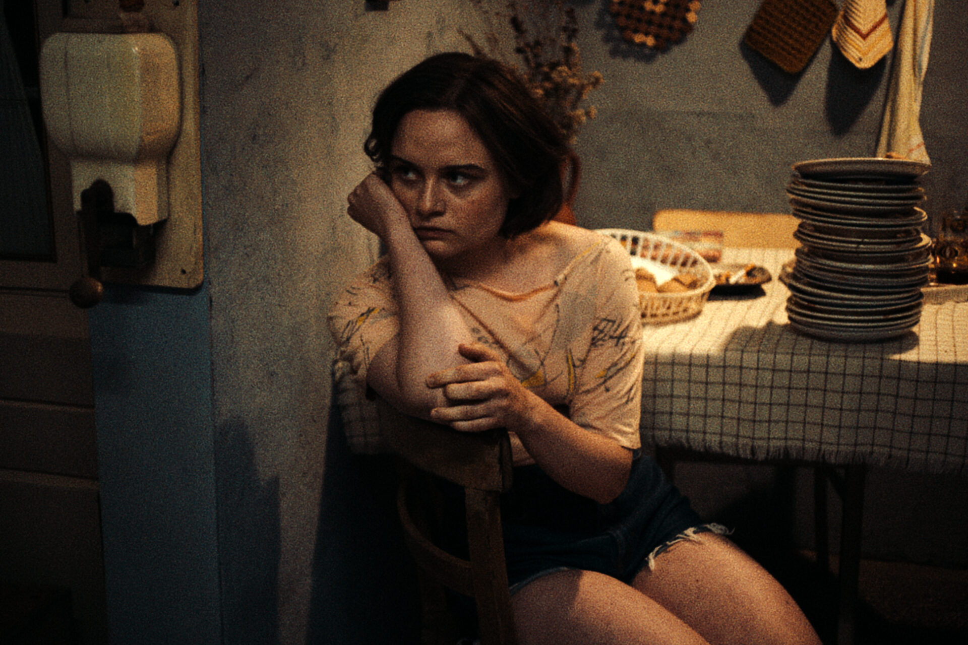 Das Bild zeigt eine Szene aus dem Film "In die Sonne schauen". Eine junge Frau sitzt auf einem Stuhl. Ihr Kopf ist auf ihrem rechten Arm gestützt.