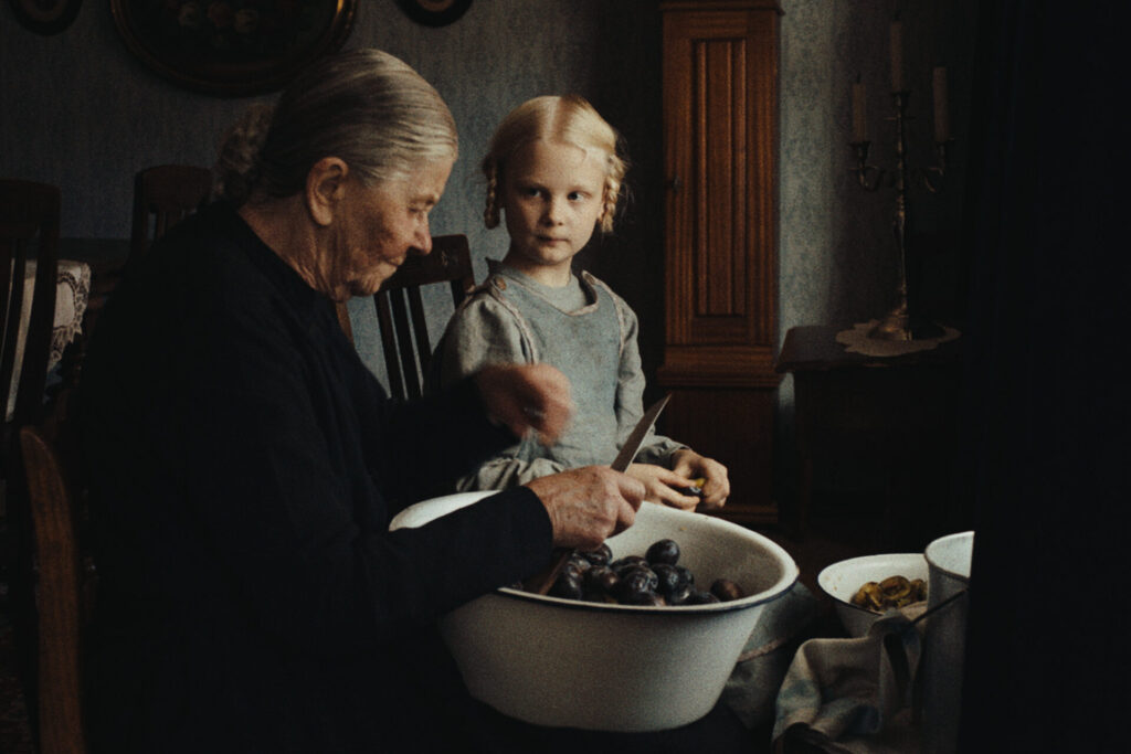 Das Bild zeigt eine Szene aus dem Film "In die Sonne schauen". Eine alte Frau sitzt auf einem Holzstuhl, auf dem Schoß eine Schüssel mit Pflaumen zum entkernen. Neben ihr sitzt ein blondes Mädchen mit Zöpfen, das ihr dabei hilft