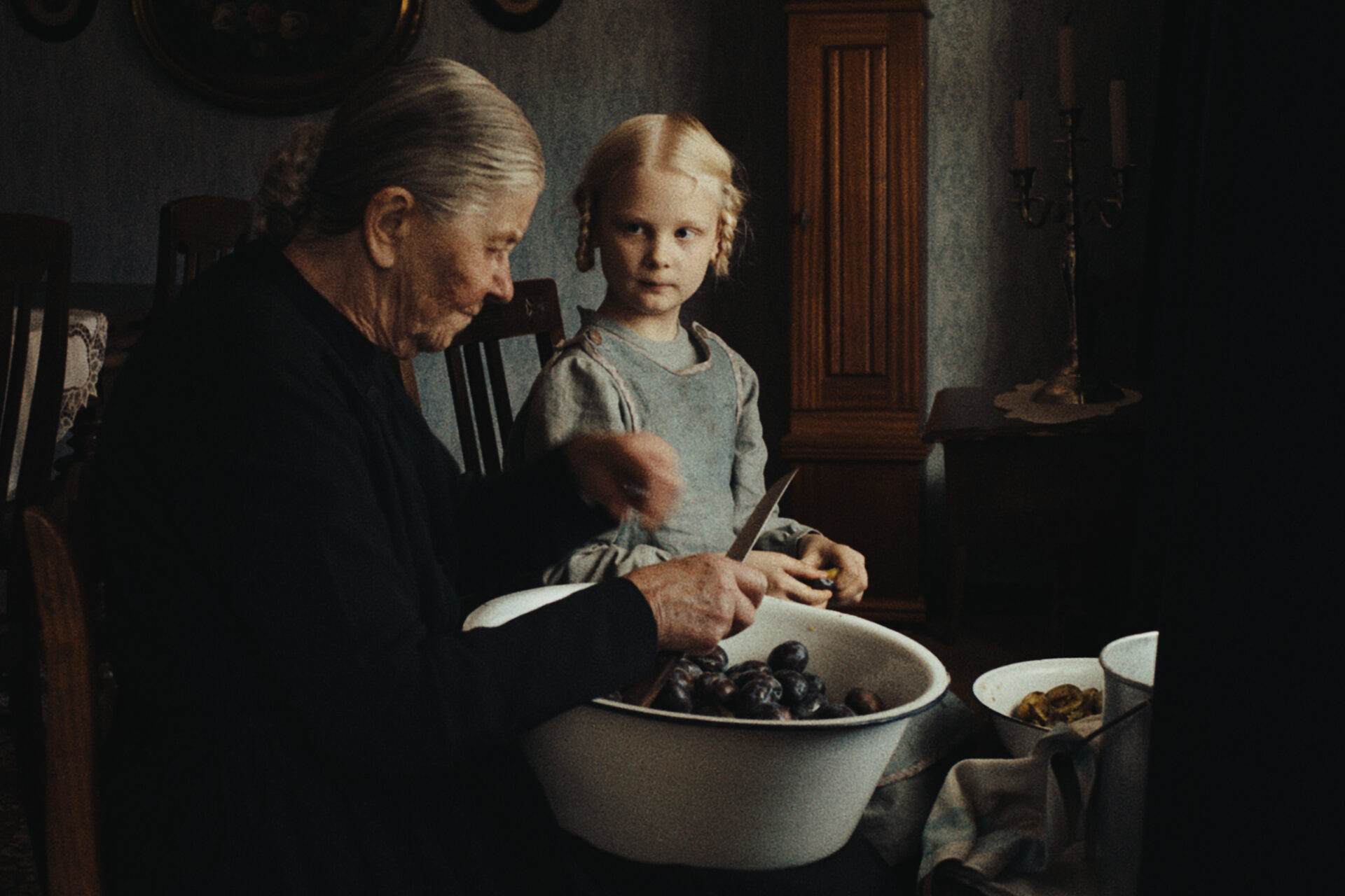 Das Bild zeigt eine Szene aus dem Film "In die Sonne schauen". Eine alte Frau sitzt auf einem Holzstuhl, auf dem Schoß eine Schüssel mit Pflaumen zum entkernen. Neben ihr sitzt ein blondes Mädchen mit Zöpfen, das ihr dabei hilft.