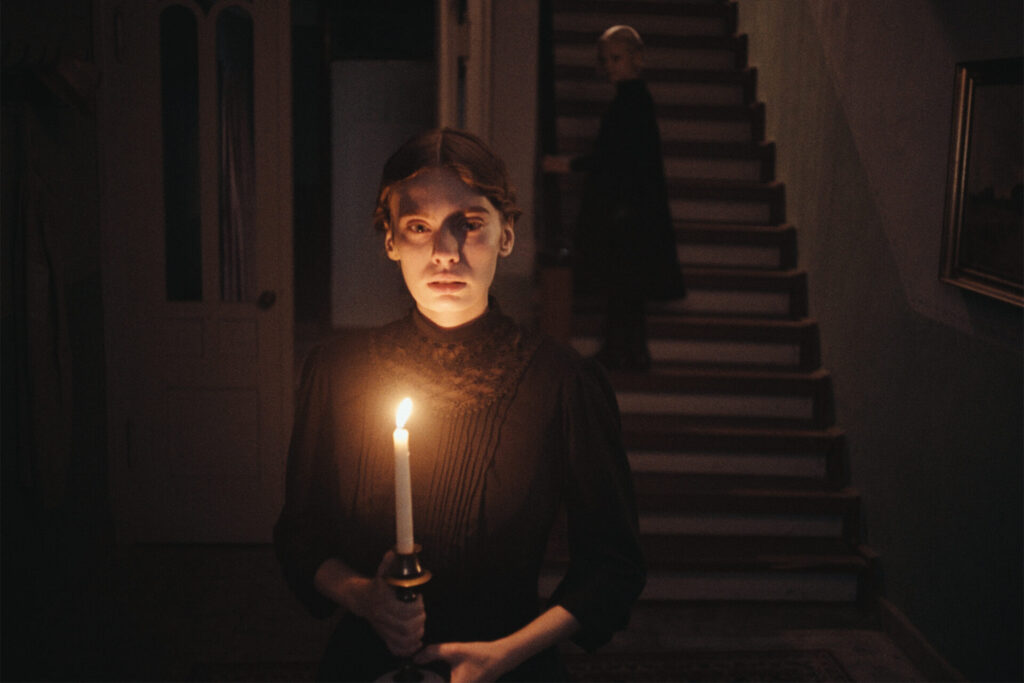 Das Bild zeigt eine Szene aus dem Film "In die Sonne schauen". Ein Mädchen steht mit einer Kerze in einem dunklen Flur. Im Hintergrund führt eine Treppe nach oben. Dort steh ein junges Mädchen.
