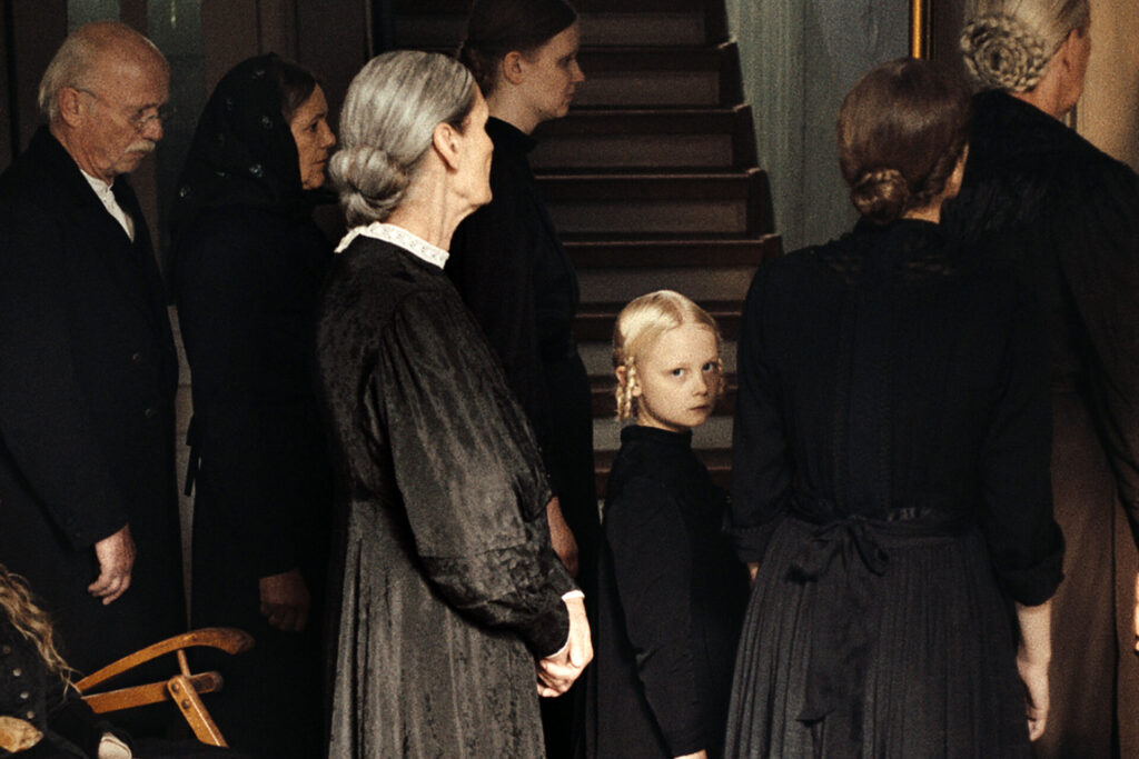 Das Bild zeigt eine Szene aus dem Film "In die Sonne schauen". Mehrere Personen in schwarzen Gewändern. Ein Mädchen schaut direkt in die Kamera. Die um sie herum stehenden erwachsenen schauen nach rechts.