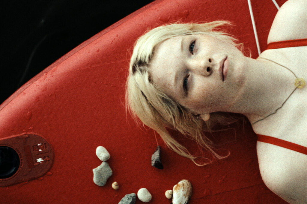 Das Bild zeigt eine Szene aus dem Film "In die Sonne schauen". Ein Mädchen liegt auf einem roten Standupboard und schaut nach oben. Sie schwimmt damit auf dem Wasser.