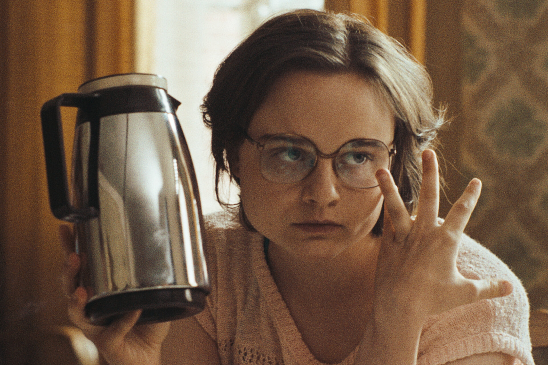 Das Bild zeigt eine Szene aus dem Film "In die Sonne schauen". Eine junge Frau hält die 3 Finger der linken Hand hoch. In der rechten Hand hat sie eine Kaffeekanne.