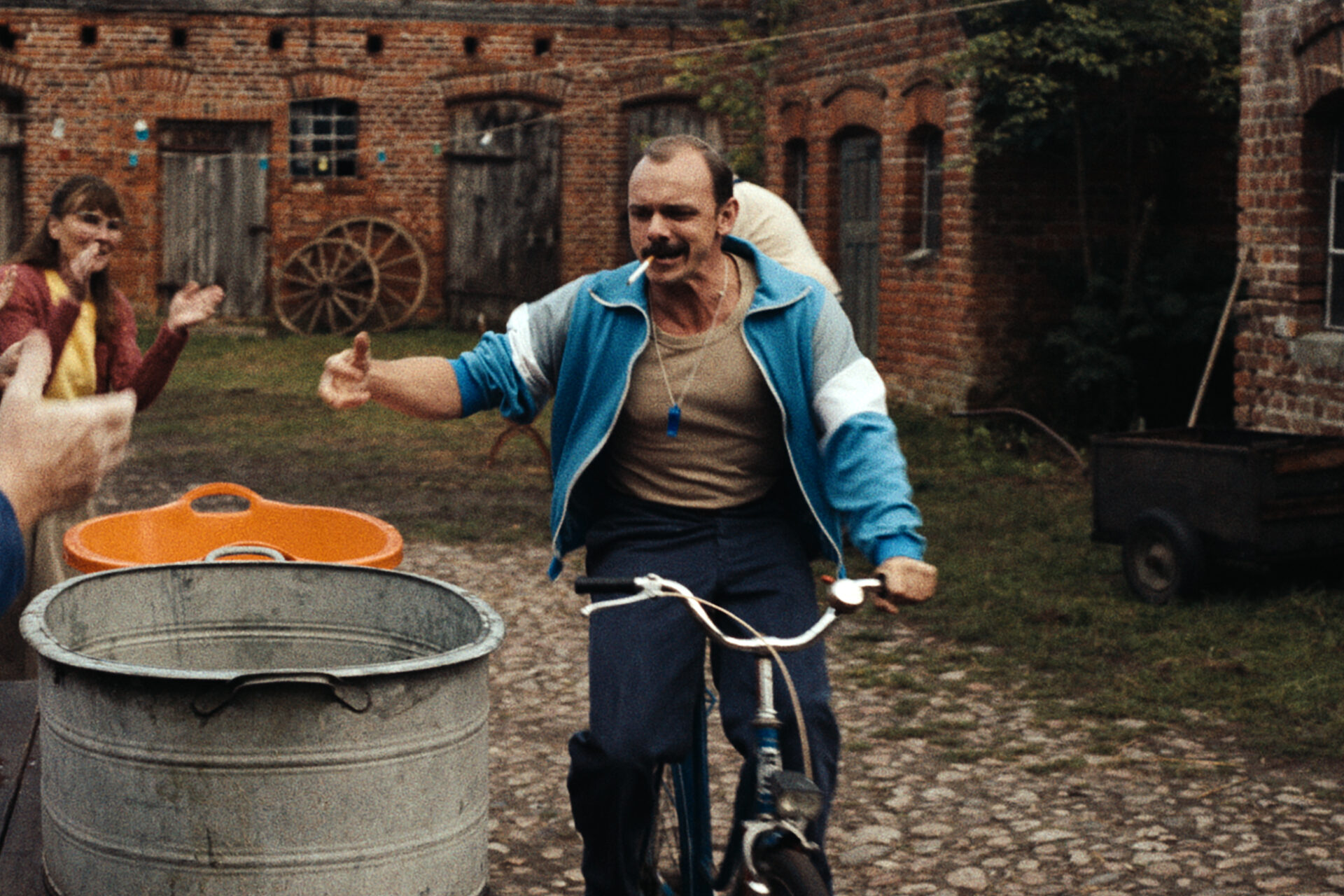 Das Bild zeigt eine Szene aus dem Film "In die Sonne schauen". Ein Mann fährt in einem Hof mit einem Fahrrad und will gerade etwas in einen Bottich werfen.