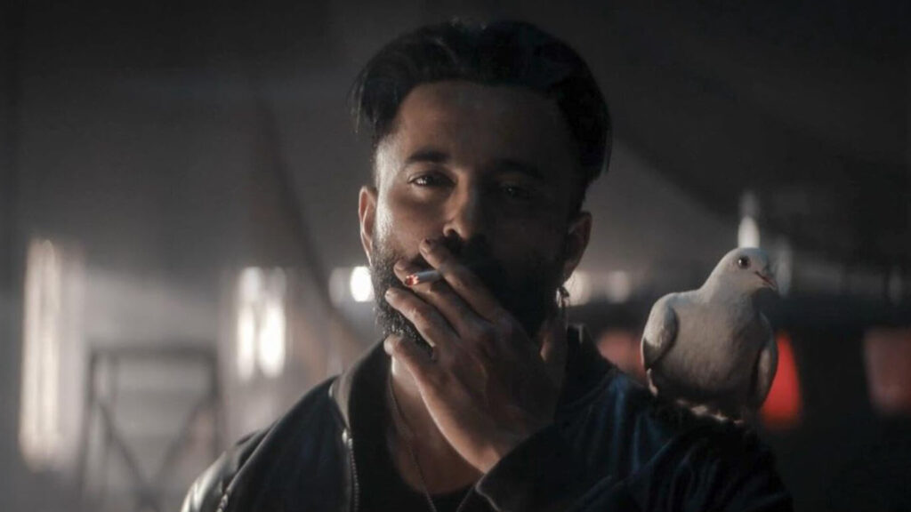 Das Bild zeigt Unni Mukundan in einer Szene des Films Marco. Er raucht und hatte eine Taube auf der Schulter.