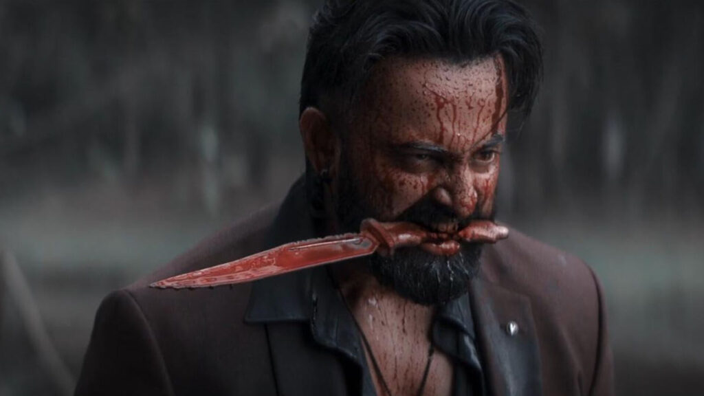 Das Bild zeigt Unni Mukundan in einer Szene des Films Marco. Er hat ein blutverschmiertes Messer im Mund,