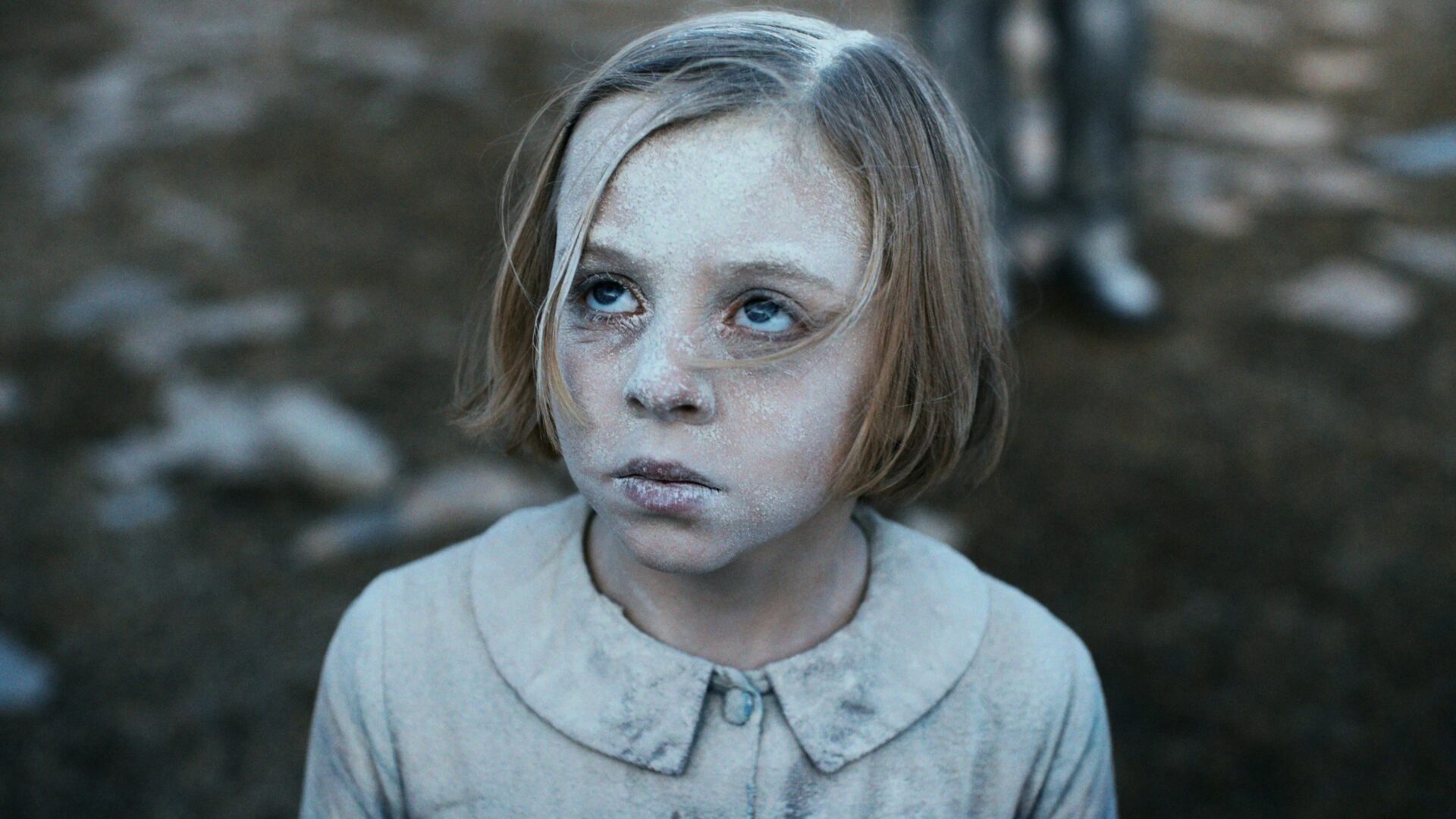 Ein kleines Kind mit kurzem blondem Haar trägt ein helles, knöpfbares Kleidungsstück. Gesicht und Kleidung sind mit einer weißen, pudrigen Substanz bedeckt, was dem Kind ein geisterhaftes, surreal wirkendes Aussehen verleiht. Der Blick des Kindes ist leicht nach oben gerichtet und wirkt leer oder nachdenklich. Der Hintergrund ist unscharf, zeigt aber eine Außenumgebung mit erdiger oder kiesiger Fläche und undeutlichen Figuren oder Objekten in der Ferne. Die Bildkomposition erzeugt eine geheimnisvolle, fast andereweltliche Stimmung durch den Kontrast zwischen dem blassen Kind und dem dunkleren Hintergrund. | 2020er Jahre Kino