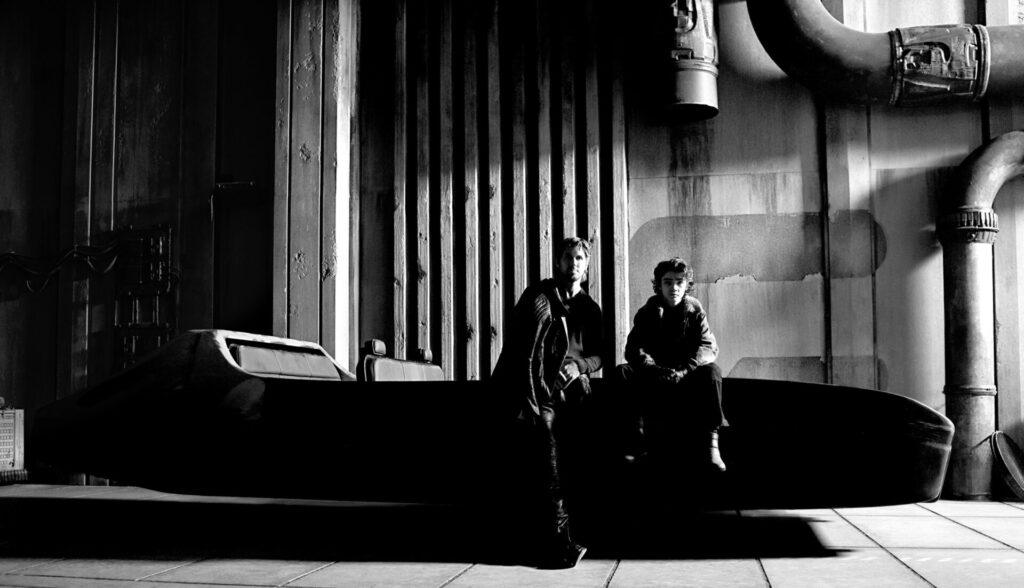 Ryan Gosling und Flynn Gray stehen und sitzen vor einer massiven, industriellen Kulisse mit großen Rohren, Lüftungsschächten und metallischen Wänden. Sie befinden sich auf oder neben einem dunklen, futuristischen Fahrzeug oder Podest. Die Szene ist dramatisch ausgeleuchtet, mit starken Licht-Schatten-Kontrasten, und vermittelt eine düstere, cineastische Atmosphäre im Stil eines dystopischen Science-Fiction-Settings. | Media Monday #741