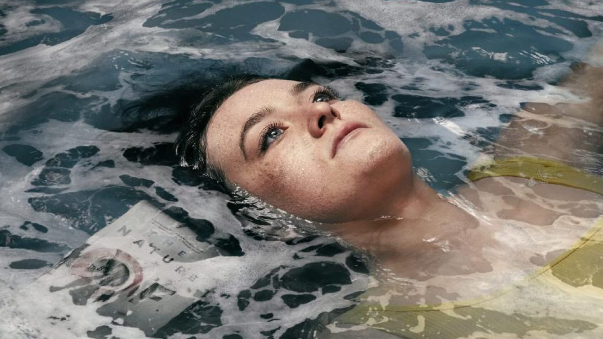 Eine Person mit dunklen Haaren und gelbem Oberteil treibt teilweise untergetaucht in aufgewühltem, schaumigem Wasser. Neben der Schulter ist ein Magazin oder Buch sichtbar. Die Szene wirkt surreal und künstlerisch, mit einem starken Kontrast zwischen Mensch und Natur. | Media Monday #741