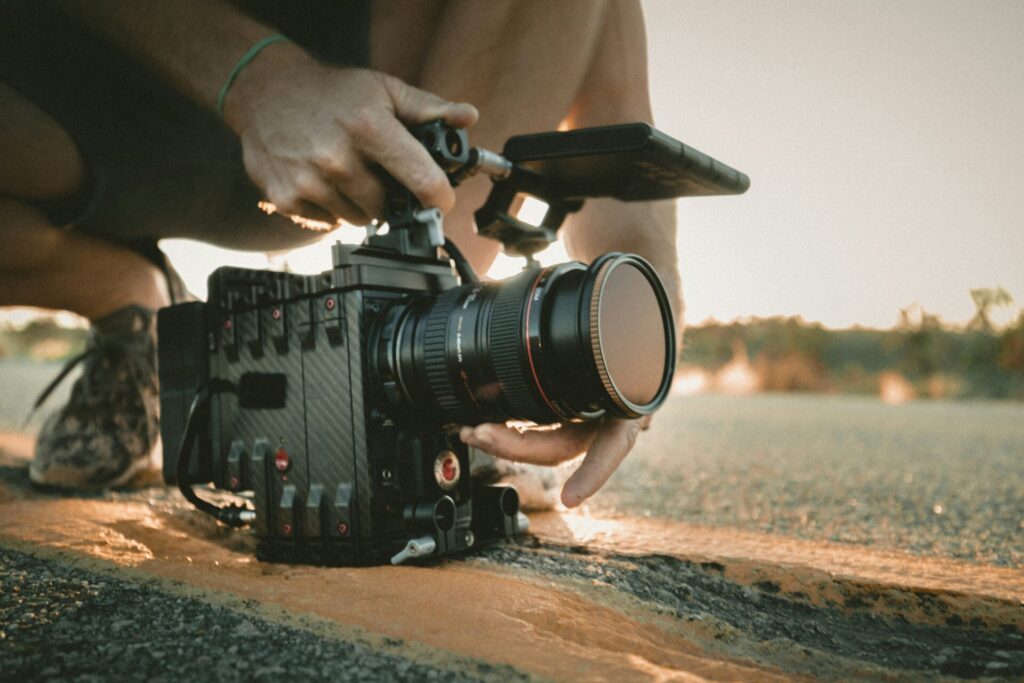 Eine Person kniet auf dem Asphalt und richtet konzentriert eine professionelle Filmkamera mit Mattebox und externem Monitor aus – ein Moment der Vorbereitung im goldenen Licht des Spätnachmittags, der die stille Hingabe hinter der Kameraarbeit sichtbar macht.