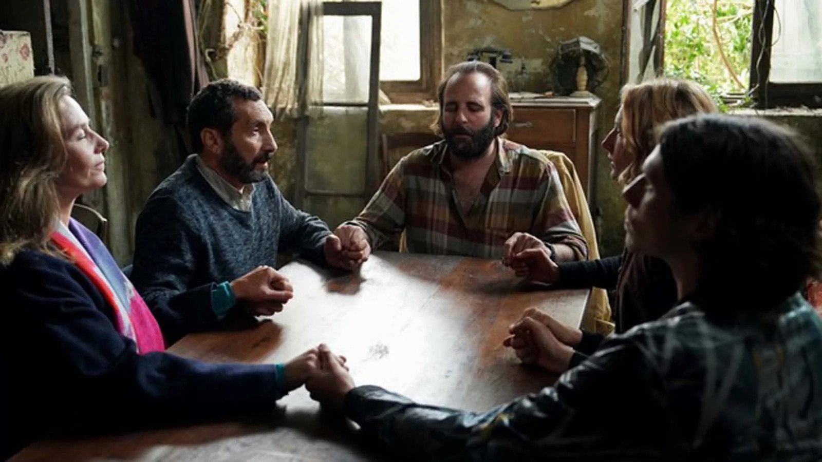 Fünf Personen sitzen an einem Tisch und halten sich an den Händen. Sie haben ihre Hände geschlossen. | Die Farben der Zeit