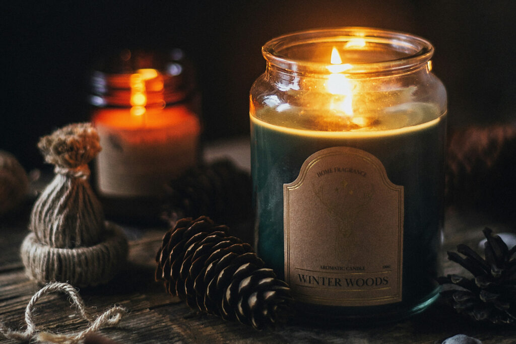 Zwei brennende Duftkerzen stehen auf einer Holzoberfläche, umgeben von Naturmaterialien wie Tannenzapfen und einem kleinen, mit Jute umwickelten Dekoelement. Im Vordergrund befindet sich eine große Glaskerze mit grünem Wachs und der Aufschrift „Winter Woods“ von der Marke „Home Purchased“. Dahinter leuchtet eine kleinere braune Kerze. Die Szene wirkt warm, rustikal und einladend – passend zur Winter- oder Feiertagssaison. | Media Monday #748