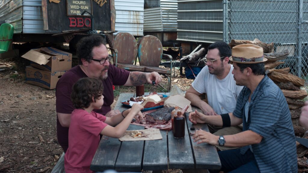 Jon Favreau und sein Sohn sitzt mit seinen Kollegen an einem Tisch im Freien und genießen ein Barbaceau | Top 5 Filme über Essen