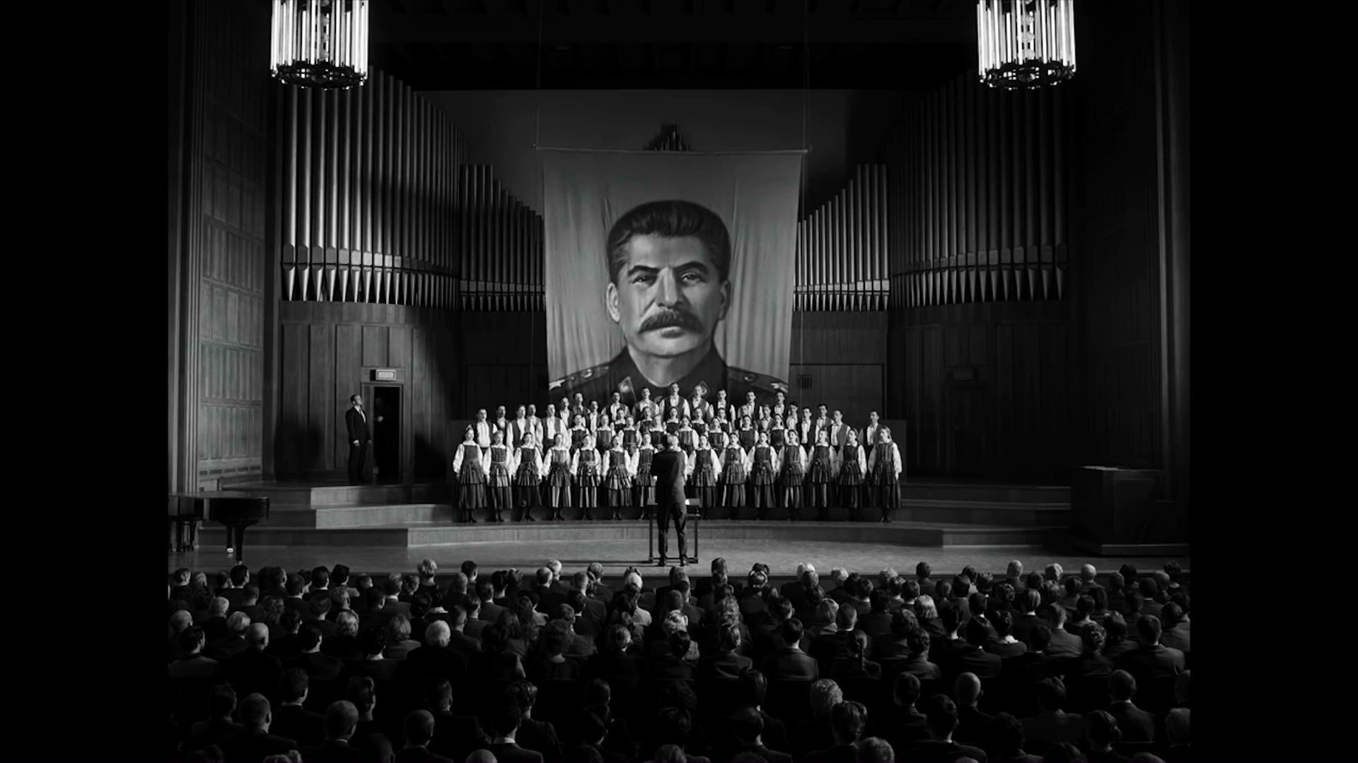 Ein Theater. auf der Bühne ist im Hintergrund ein großes Bild von Stalin zu sehen. Davor ein Chor und vor der Bühne Publikum. | Monochrome Meisterwerke: 5 Arthouse Filme in Schwarz-Weiß
