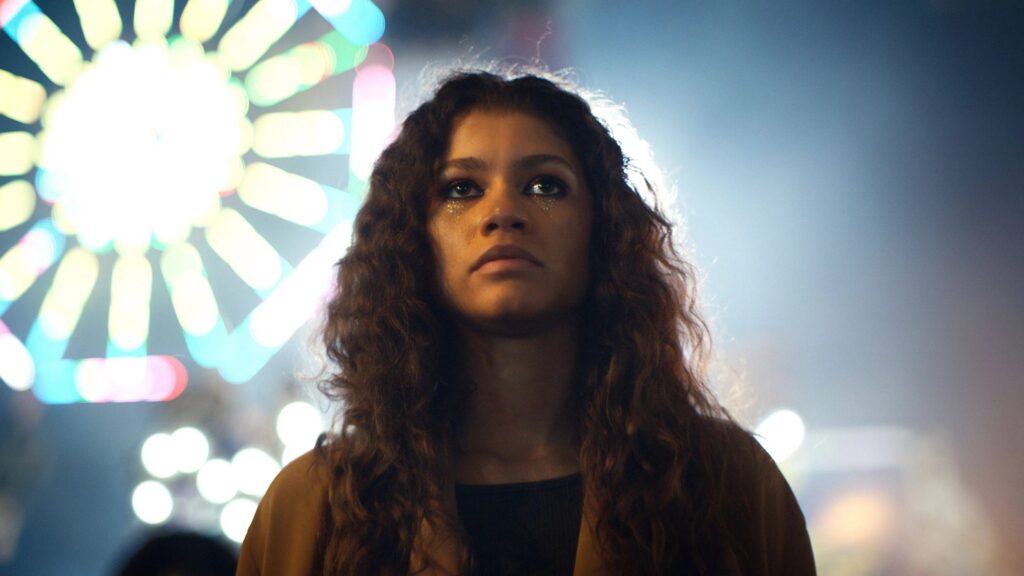 Das Bild zeigt Rue (Zendaya) aus der Serie "Euphoria" mit glitzernden Make-Up umgeben von Licht, auf dem Rummelplatz. | Themenübersicht März 2026