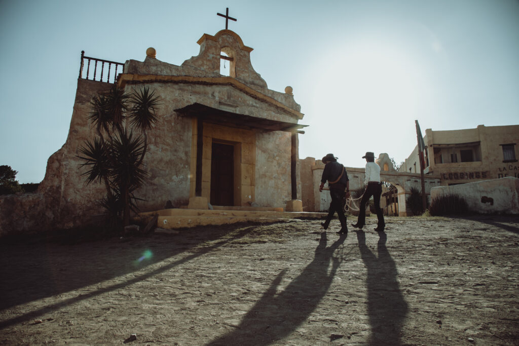 Das Bild zeigt zwei Personen in Wild-West-Kleidung vor einer Kirche in einer Szene des Films Make Me Feel