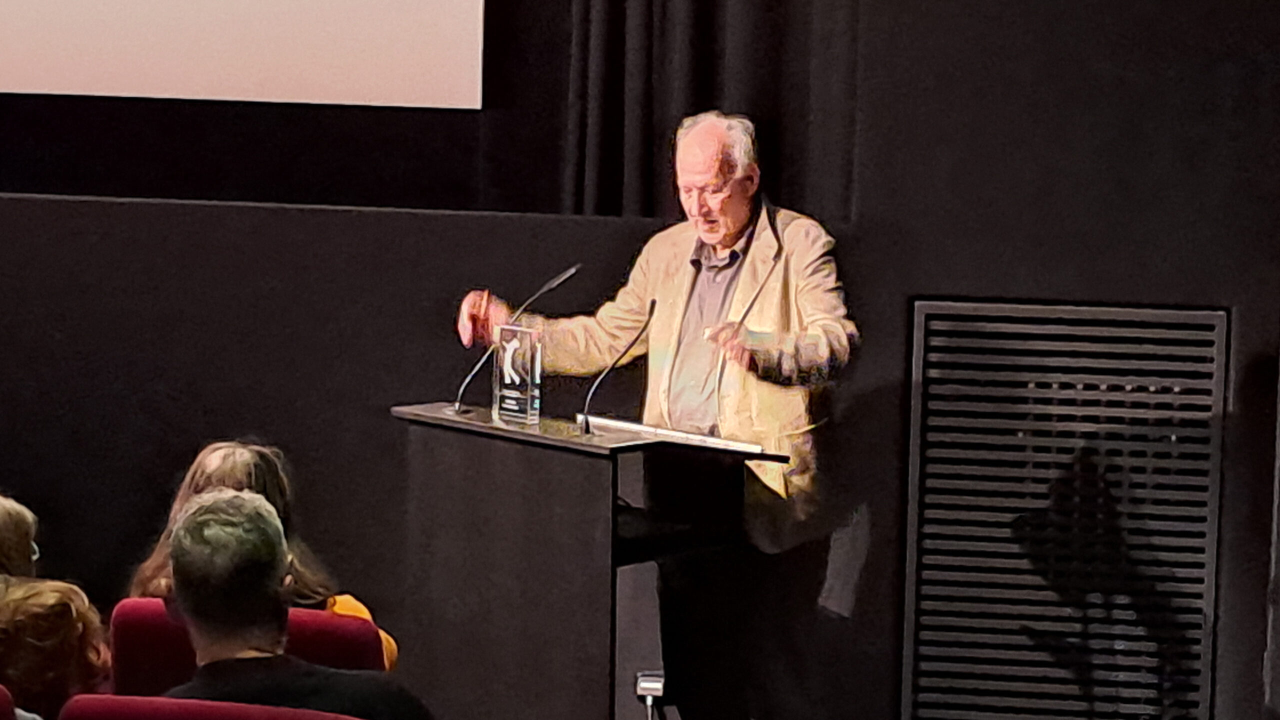 Das Bild zeigt Werner Herzog am Redner:innenpult im Filmmuseum München