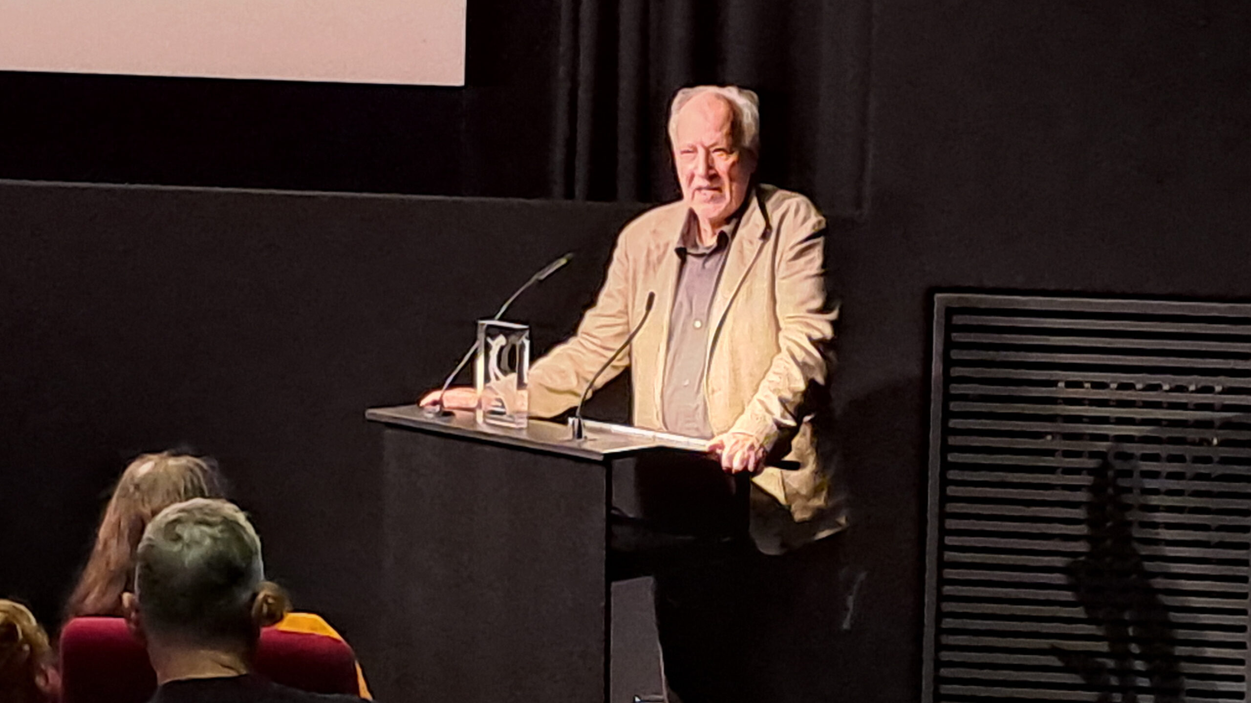 Das Bild zeigt Werner Herzog am Redner:innenpult im Filmmuseum München