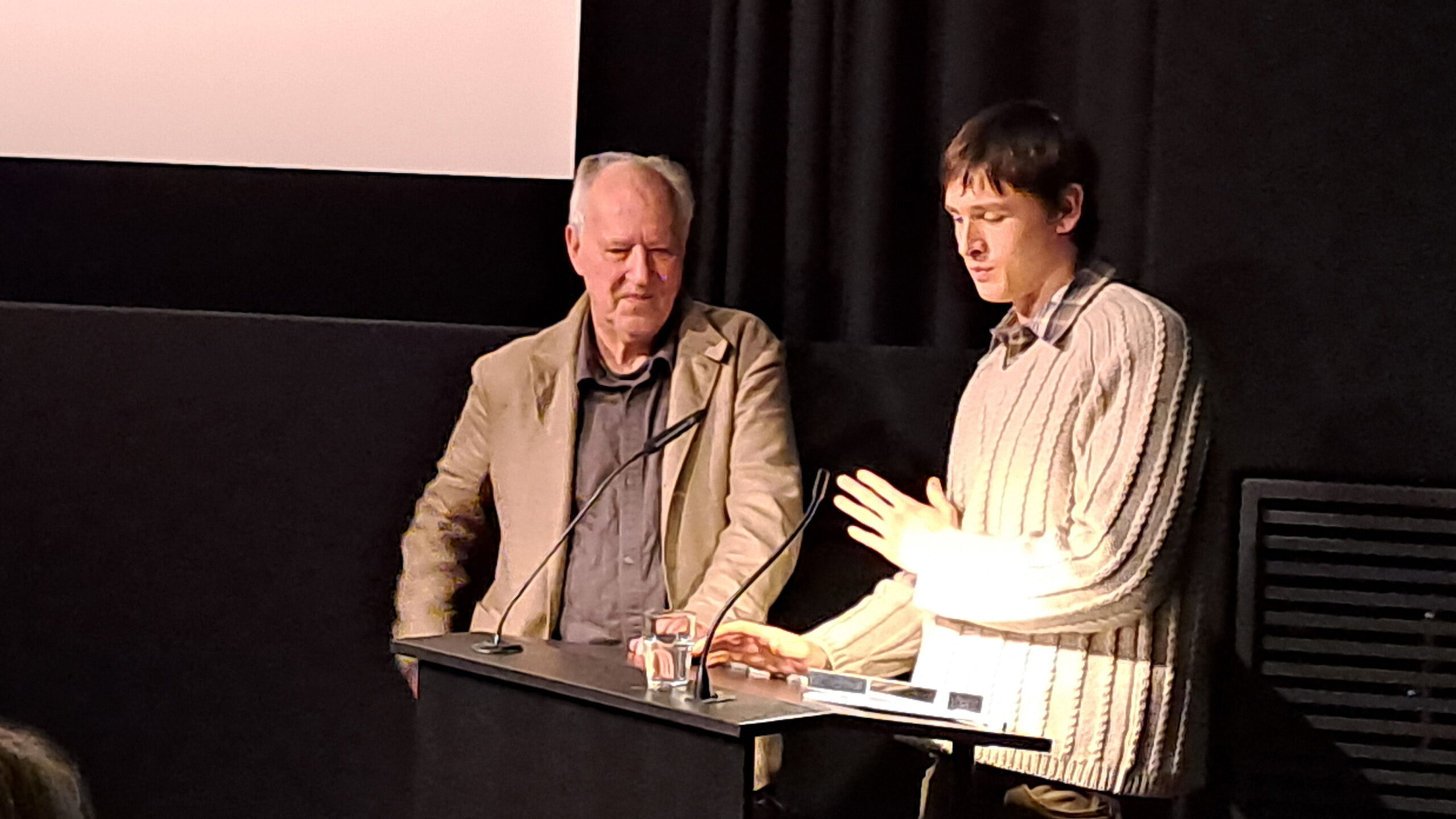 Das Bild zeigt Werner Herzog und Harris Dickinson im Filmmuseum München. Werner Herzog überreicht Dickinson gerade den Werner-Herzog-Filmpreis 2025