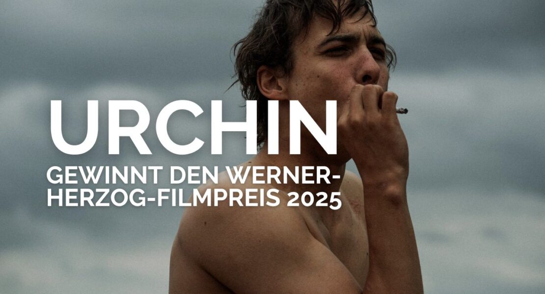 Schauspieler Frank Dillane steht oberkörperfrei vor einem wolkenverhangenen Himmel und raucht eine Zigarette. Über dem Bild ist in weißer, fetter Schrift zu lesen: „URCHIN gewinnt den Werner-Herzog-Filmpreis 2025“. Die Szene wirkt rau und atmosphärisch und kündigt den preisgekrönten Film „Urchin“ an.
