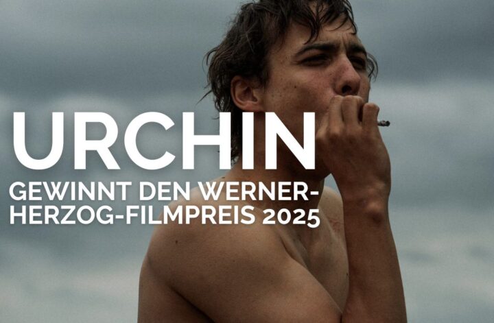 Schauspieler Frank Dillane steht oberkörperfrei vor einem wolkenverhangenen Himmel und raucht eine Zigarette. Über dem Bild ist in weißer, fetter Schrift zu lesen: „URCHIN gewinnt den Werner-Herzog-Filmpreis 2025“. Die Szene wirkt rau und atmosphärisch und kündigt den preisgekrönten Film „Urchin“ an.