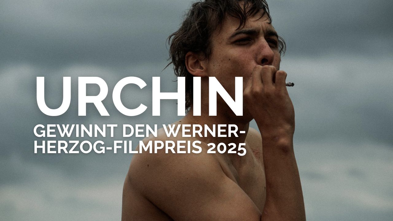 Schauspieler Frank Dillane steht oberkörperfrei vor einem wolkenverhangenen Himmel und raucht eine Zigarette. Über dem Bild ist in weißer, fetter Schrift zu lesen: „URCHIN gewinnt den Werner-Herzog-Filmpreis 2025“. Die Szene wirkt rau und atmosphärisch und kündigt den preisgekrönten Film „Urchin“ an.