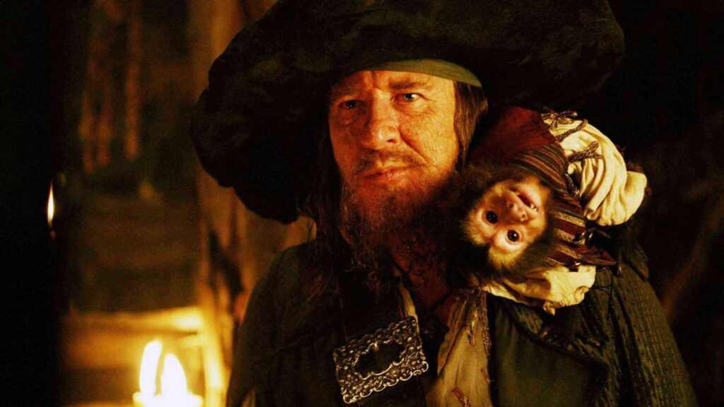 Geoffrey Rush als Captain Barbossa aus Fluch der Karibik steht in dunkler Piratenkleidung mit großem Hut im warm beleuchteten Schiffsinneren, während sein kleiner Affe Jack in passender Mini-Piratenkleidung auf seiner Schulter sitzt. | Themenübersicht März 2026