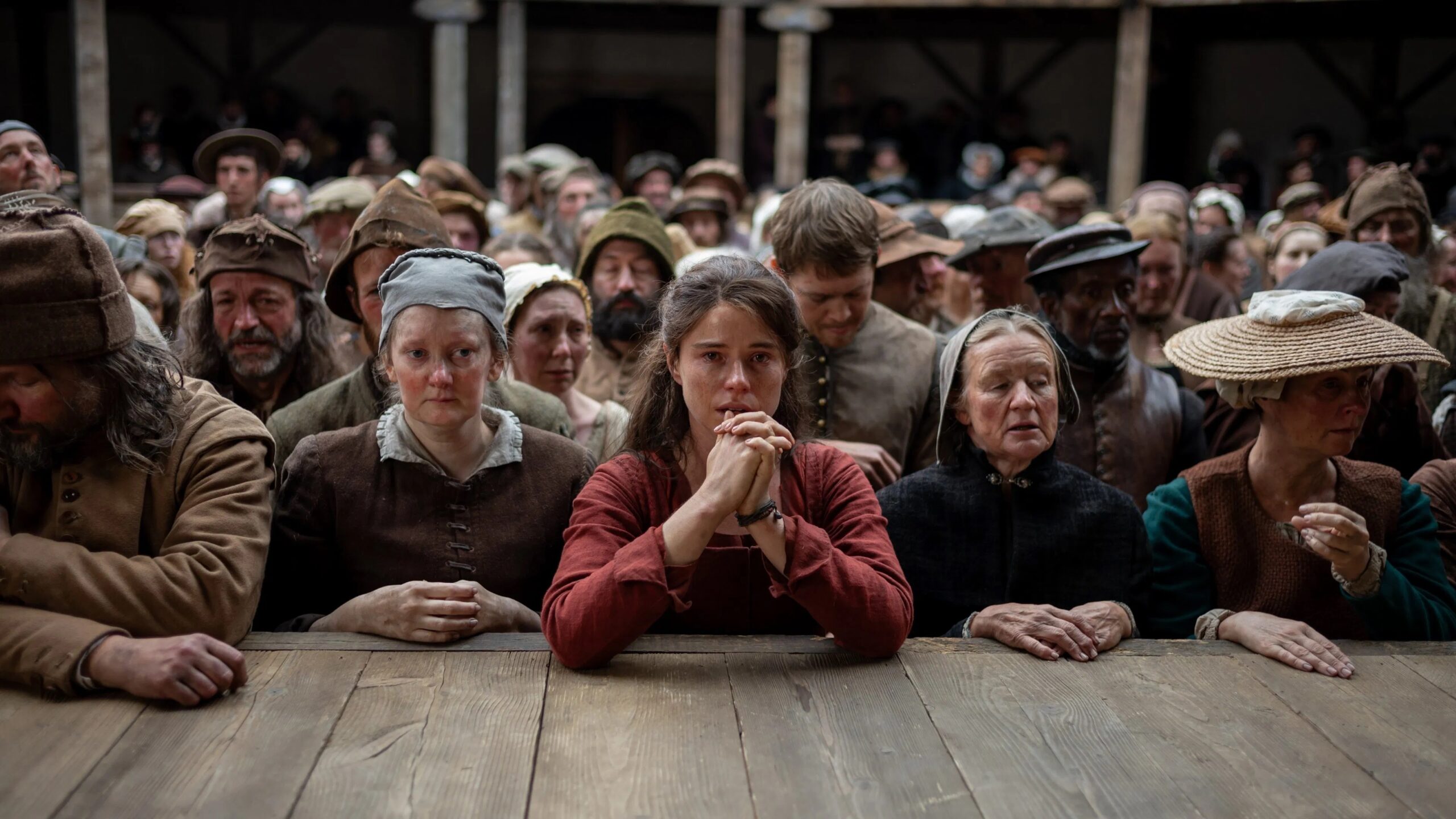 Das Bild zeigt Agnes (Jessie Buckley) mit gefalteten Händen und Tränen in den Augen am Bühnenrand stehen, die Arme auf die Bühne gestützt. Sie ist umgeben vom Rest des Publikums, das ergriffen von dem Stück ist