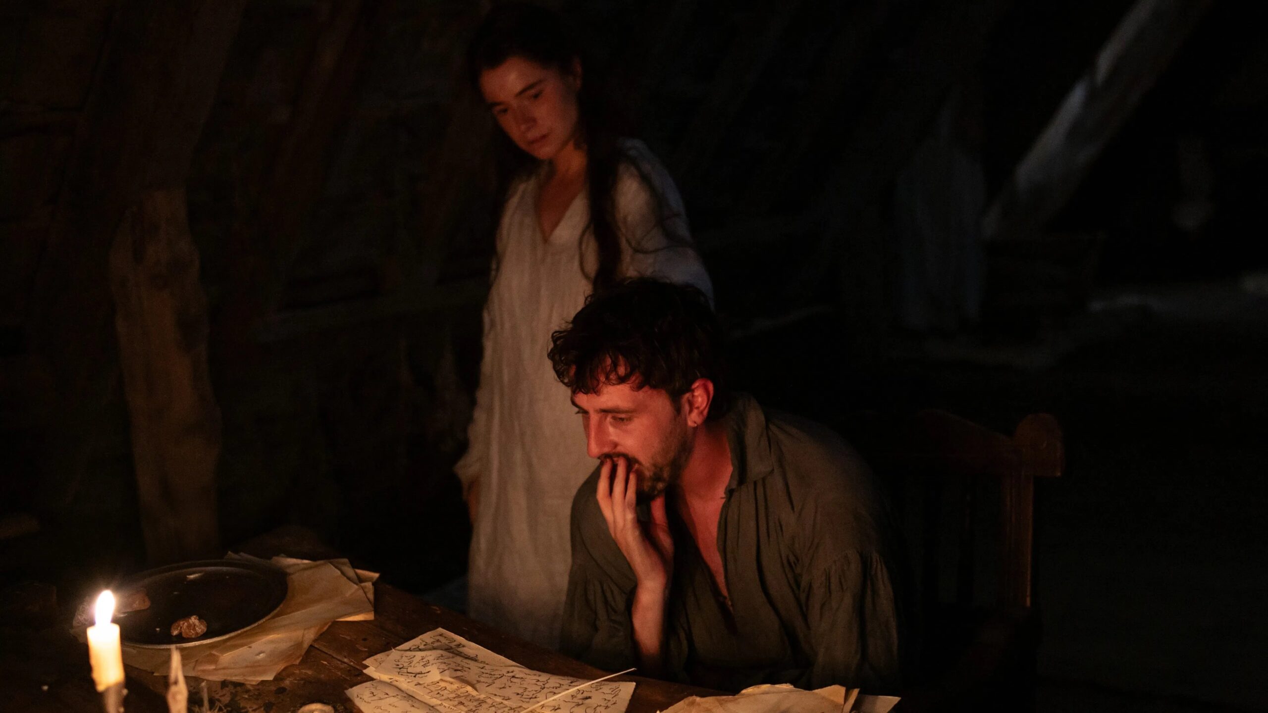 Das Bild zeigt William Shakespeare (Paul Mescal) am Schreibtisch. Hinter ihm steht seine Frau Agnes (Jessie Buckley). Dies ist eine Szene aus dem Film Hamnet von Chloé Zhao