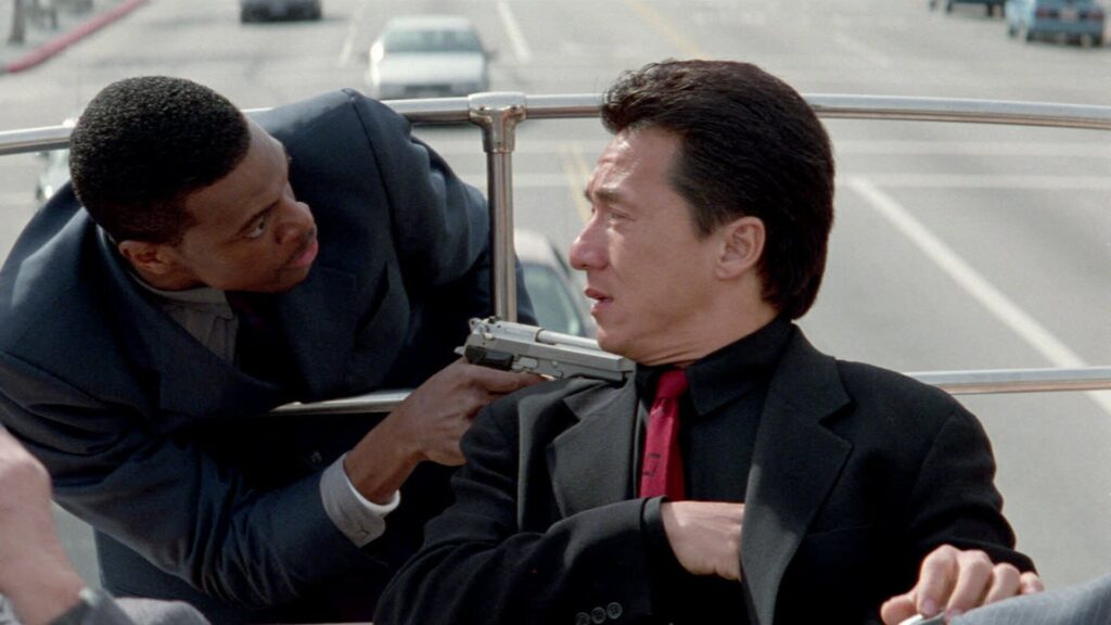 Jackie Chan sitzt an einem Geländer, Chris Tucker lehnt sich von hinten durch und zielt mit einer Pistole auf Jackie