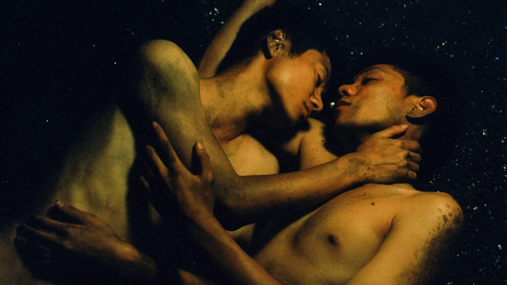 Das Bild zegt Nam (Phạm Thanh Hải) und Viet (Đào Duy Bảo Định) eng umschlungen, nackt auf dem schwarz funkelnden Boden im Kohlebergwerk. Die Szene stammt aus dem Film Viet und Nam