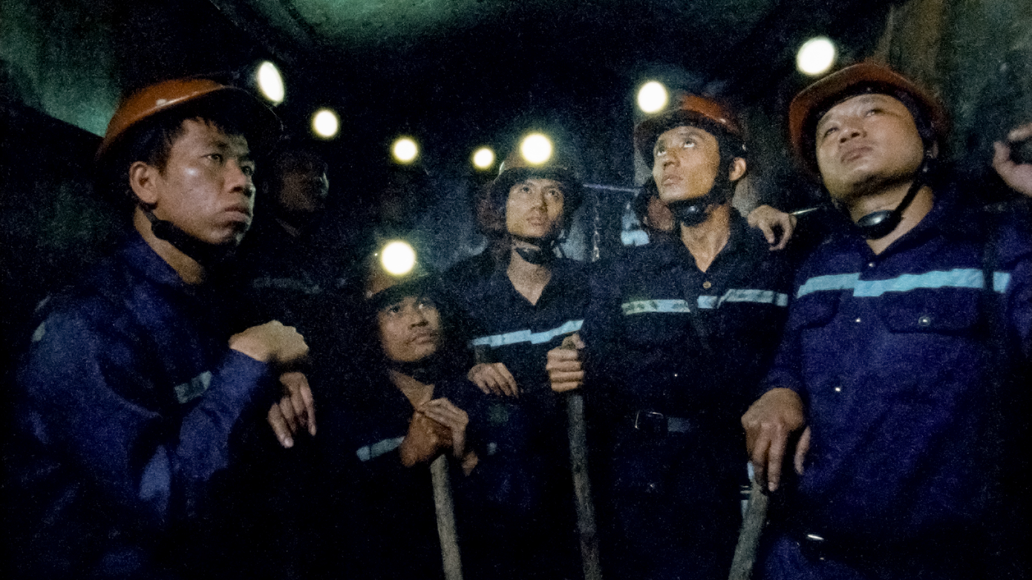 Das Bild zeigt die Bergleute auf dem Weg in den Abgrund im Aufzug stehend. Ausgerüstet mit Spitzharke, Helm mit Stirnleuchte und Overall