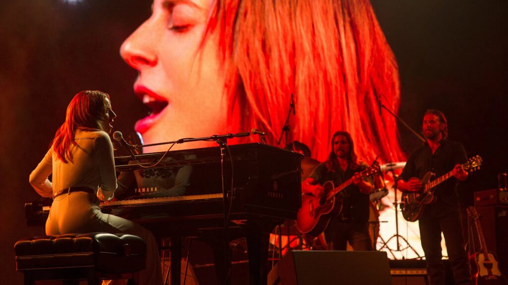 Lady Gaga sitzt an einem Klavier, während zwei Männer im Hintergrund Gitarre spielen, dahinter ist das Gesicht von Lady Gaga auf einer großen Leinwand zu sehen