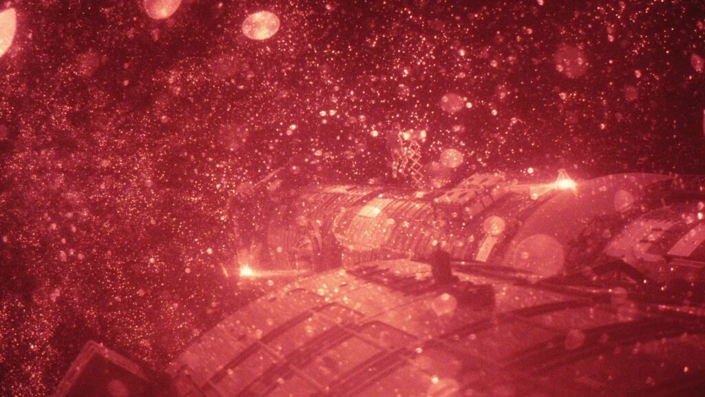 Rötlich beleuchteter Ausschnitt der Raumsonde Hail Mary aus Der Astronaut. Im Vordergrund ist ein zylindrisches Modul mit technischen Paneelen zu sehen. In der Ferne, sehr klein, schwebt die Figur von Ryland Grace (Ryan Gosling). Rundherum verteilt sich ein dichter Schwarm heller Partikel, der wie schwebender Staub oder Sternengestöber wirkt und dem Bild Tiefe verleiht. | Themenübersicht April 2026