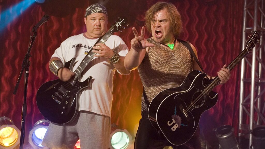 Jack Black und Kyle Gass stehen auf einer Bühne und haben jeweils eine Gitarre in der Hand. Um den Hals von Jack Black hängt ein leuchtends Plektrum, während er singt. | Die 5 besten Musik-Biopics