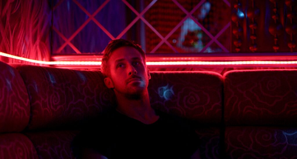 Julian (Ryan Gosling) sitzt in einem schwach beleuchteten Raum, der in intensives Rot‑ und Violettlicht getaucht ist. Er sitzt auf einem gemusterten Sofa, umgeben von metallischen Strukturen und hängenden Dekorationen, die das Licht reflektieren. Die Szene wirkt still, angespannt und fast traumartig, typisch für die stilisierte Atmosphäre von Only God Forgives. | Themenübersicht April 2026