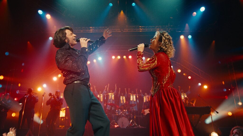 Hugh Jackman und Kate Hudson im Film "Song Sung Blue" stehen sich auf einer Bühne gegenüber und singen, während im Hintergrund die Band und ein Chor zu sehen ist | Die 5 besten Musik-Biopics