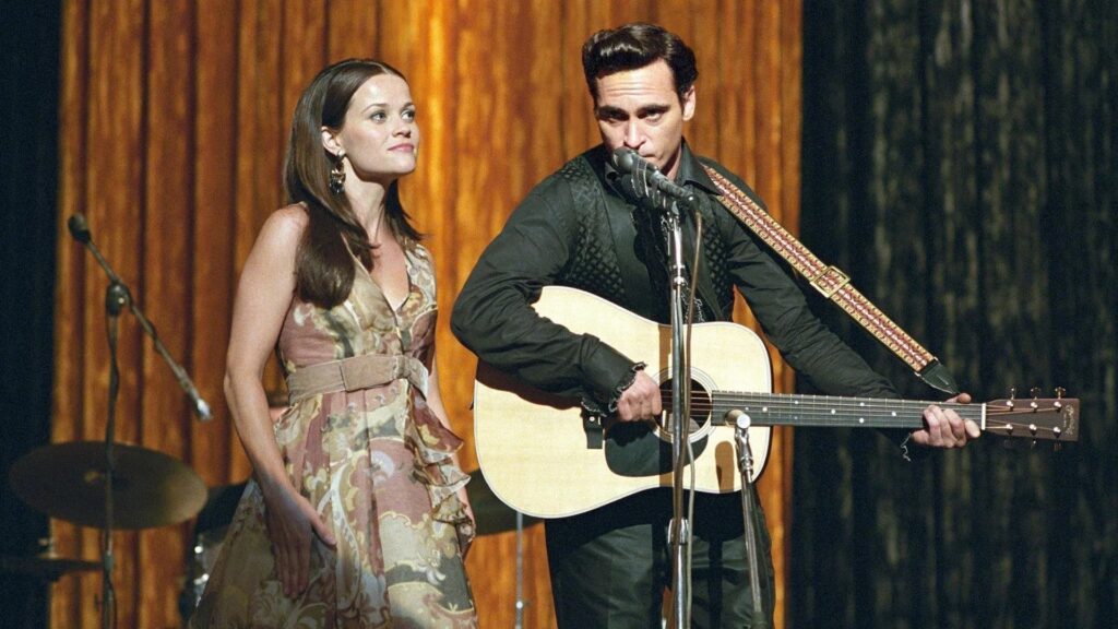 Joaquin Phoenix als Johnny Cash und Reese Witherspoon als June Carter stehen gemeinsam auf einer Bühne, er hält eine Gitarre im Arm und spielt | Die 5 besten Musik-Biopics
