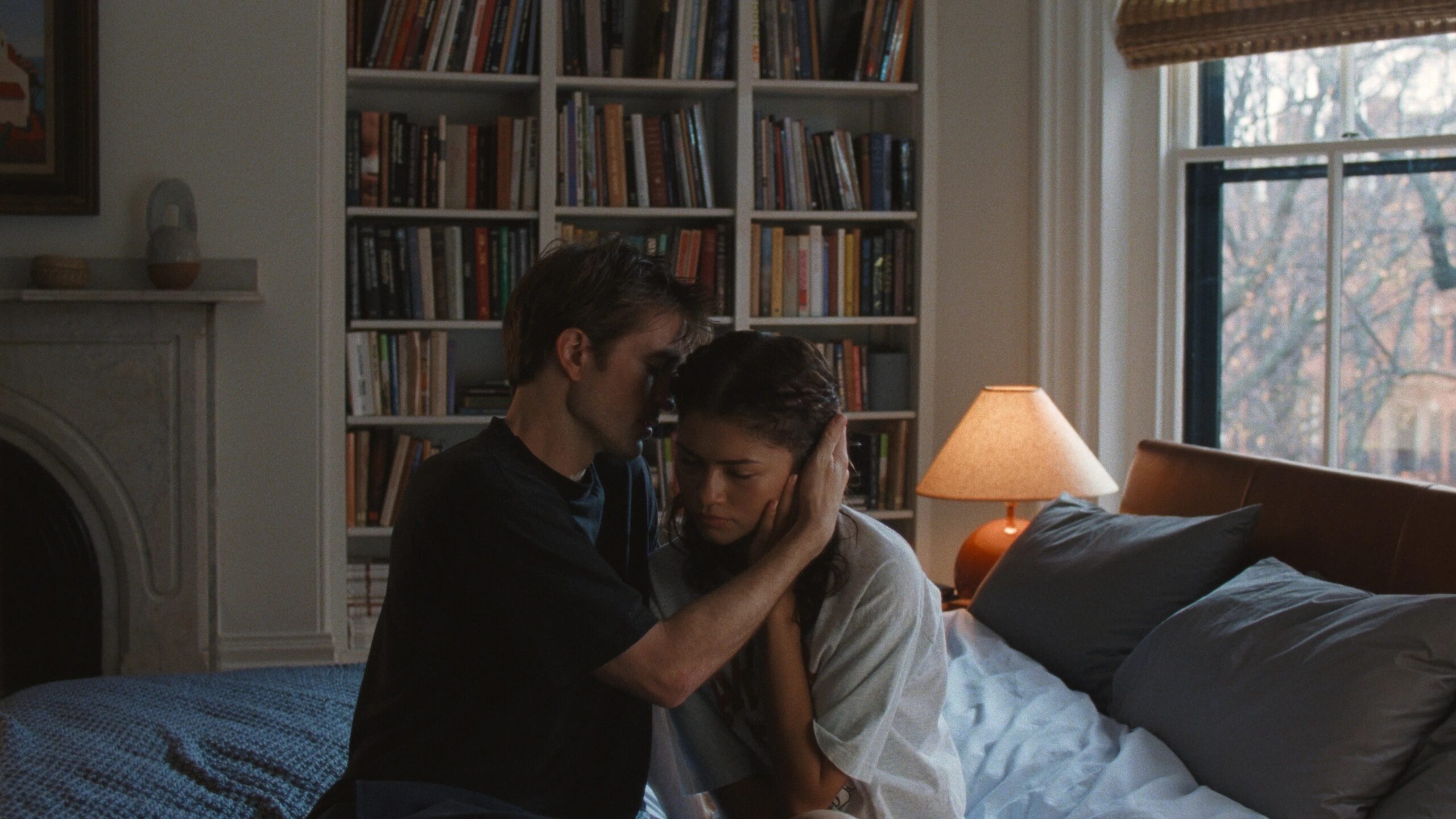 Zendaya und Robert Pattinson in einer intimen Szene aus Das Drama: Sie sitzen eng beieinander auf einem Bett, eine beruhigende Geste, warmes Licht und Bücherregal im Hintergrund.