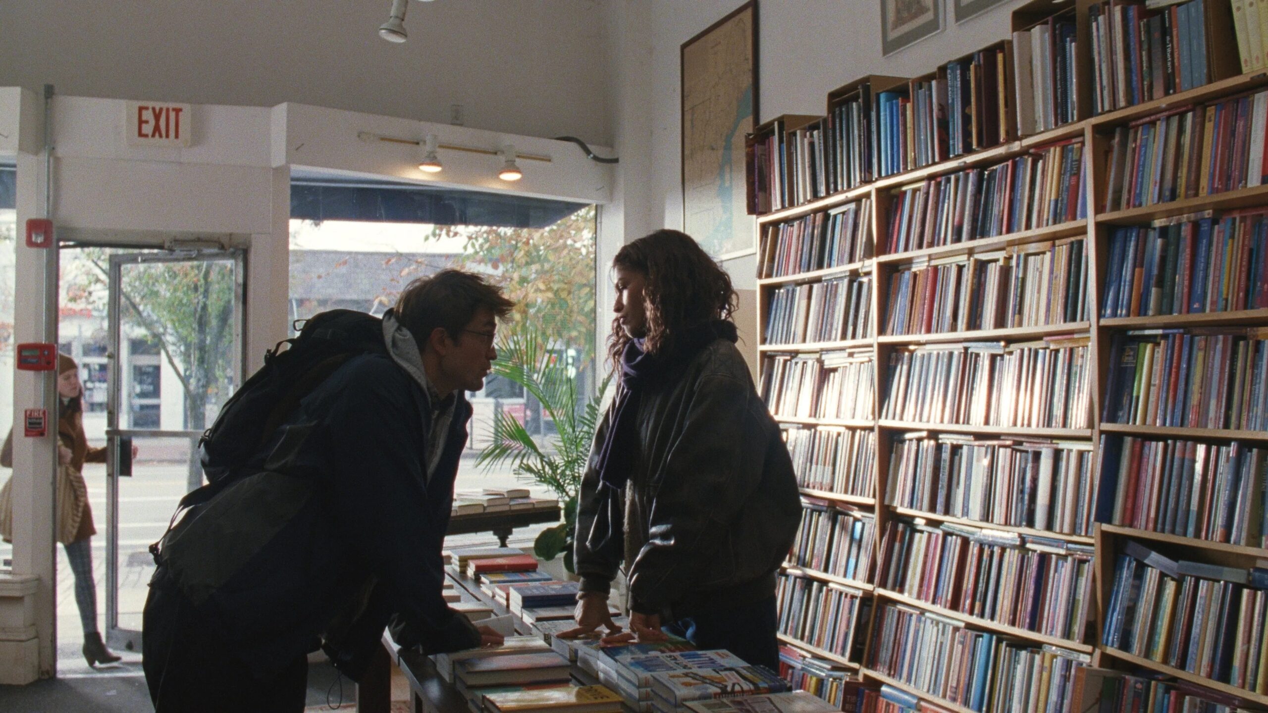 Zendaya und Robert Pattinson in einer Szene aus Das Drama in einer Buchhandlung, im Gespräch zwischen hohen Bücherregalen.