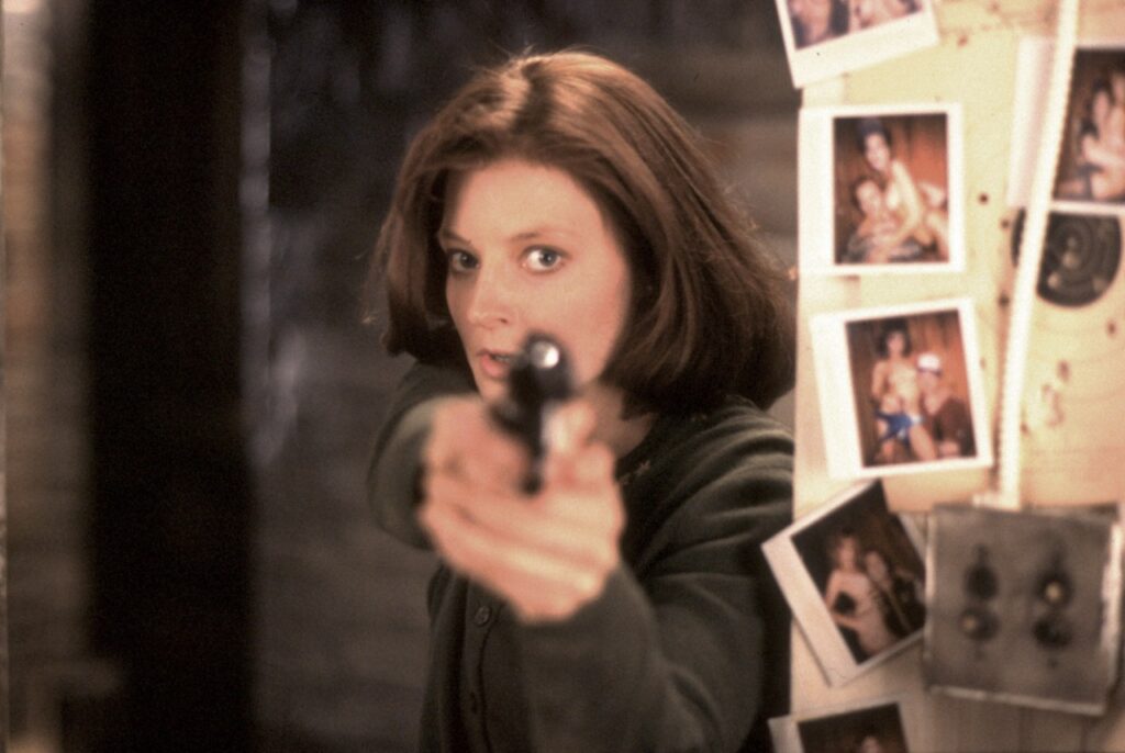 Jodie Foster als Clarice Starling zielt mit einer Pistole in einem dunklen Raum; im Hintergrund hängen Polaroid‑Fotos an einer Wand – ikonische Szene aus ‚Das Schweigen der Lämmer‘.