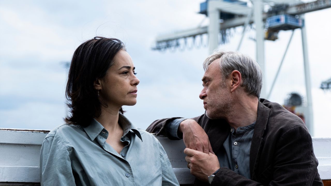 Eine Frau und ein Mann sitzen an einem Hafen. Im Hintergrund ist ein großer Lastkran zu sehen.