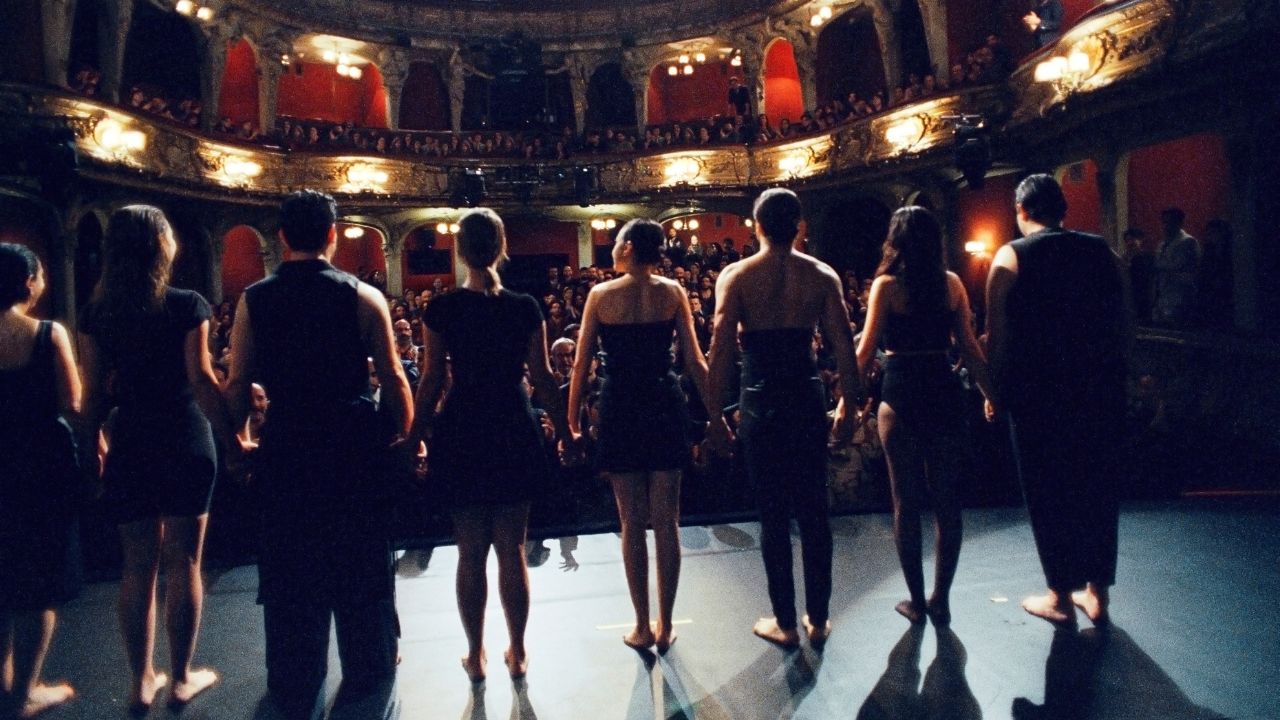 Mehrere Menschen stehen auf einer Theaterbühne mit dem Rücken zur Kamera. In ihrer Blickrichtung ist der gefüllte Zuschauerraum. | Gelbe Briefe