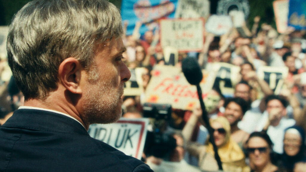 Ein Mann steht vor einem Mikrofon. Im Hintergrund (in seiner Blickrichtung) steht eine Menge, welche Plakate und Transparente in die Höhe hält. | Gelbe Briefe Filmkritik