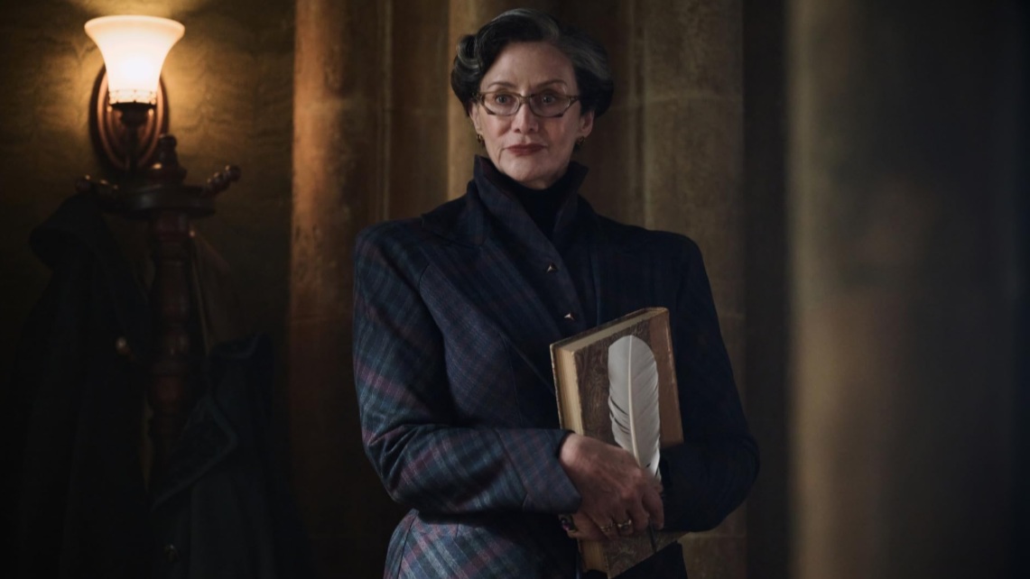 Minerva McGonagell in der Harry Potter Serie steht in einem Gang und hält ein Buch mit einer Feder an ihren Körper gepresst.