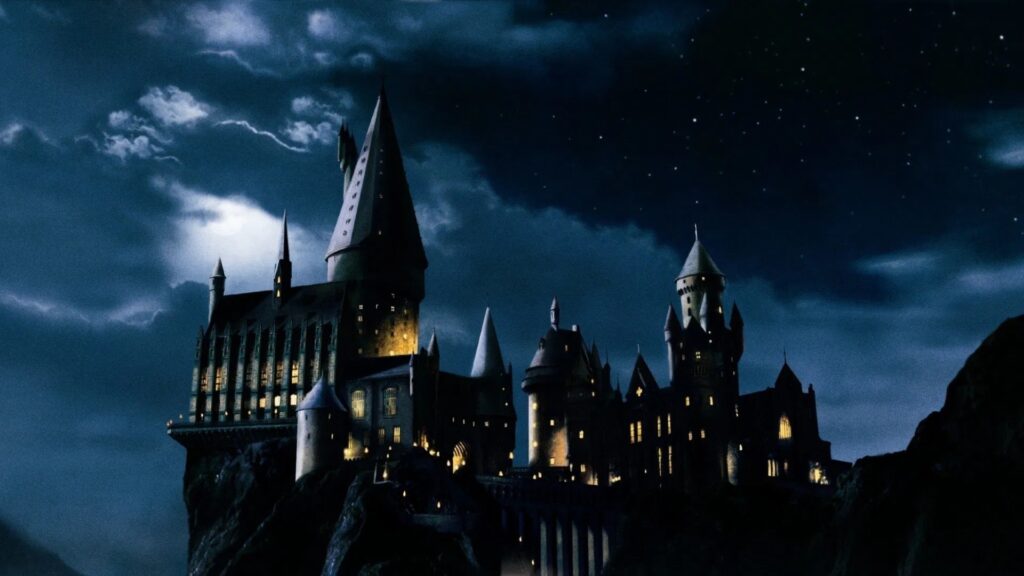 Ein Panorama des Schlosses Hogwarts bei Nacht, der Himmel ist bewölkt, die Fenster und Tore des Schlosses beleuchtet