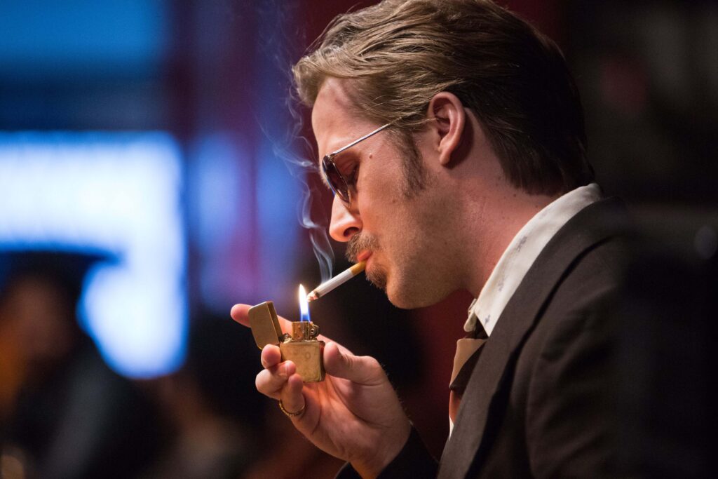 Ryan Gosling als Holland March in The Nice Guys, im Anzug mit Sonnenbrille, zündet sich eine Zigarette an; stilvolle, rauchige Neo‑Noir‑Atmosphäre. | Media Monday #774
