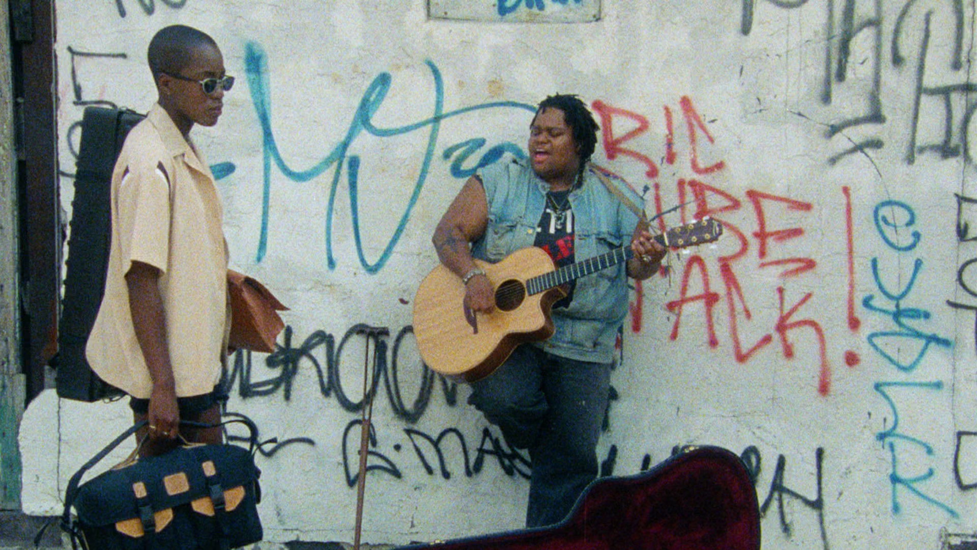 Eine Person mit Gitarre lehnt an einer graffitibesprühten Wand und singt. Links steht Cheryl mit einer Tasche in der Hand.